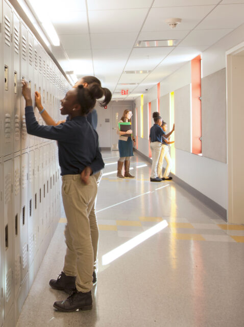 Students unlocking grey lockers in sun lit corrdior lined with windows accented by yellow and orange.