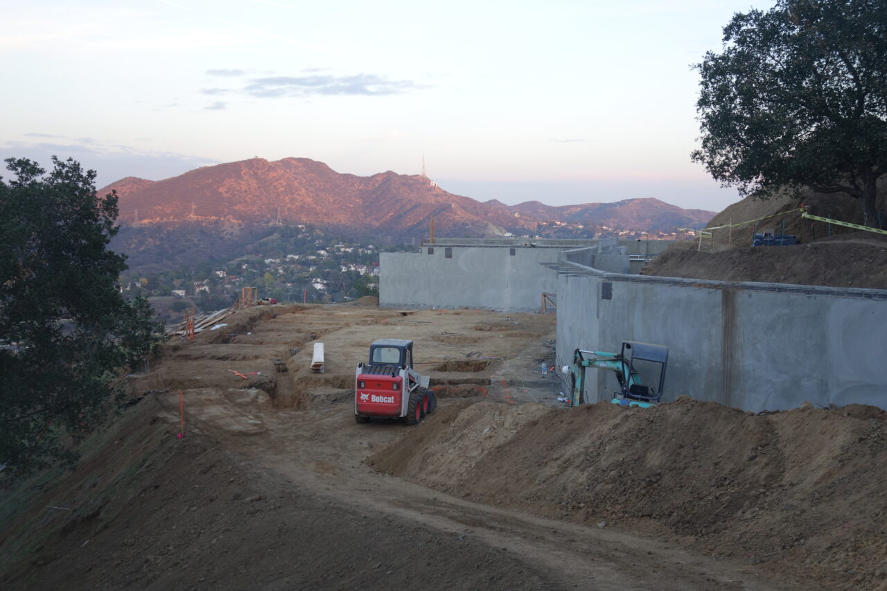 Excavated site held by concrete retaining wall framed by vegetation and hills in background.